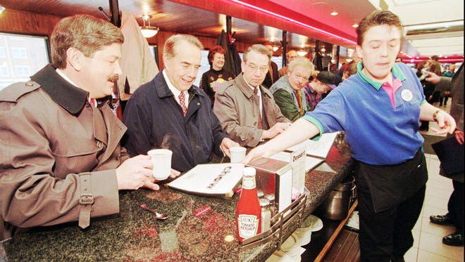 Waitress Chris Cook, right, closes her eyes to the bright TV lights, as she slides a menu in front of Iowa Gov. Terry Branstad, left, Kansas Sen. and presidential candidate Bob Dole and Iowa Sen. Charles Grassley, during a campaign visit to the Drake Diner in Des Moines, Iowa, Sunday, Feb. 11, 1996.